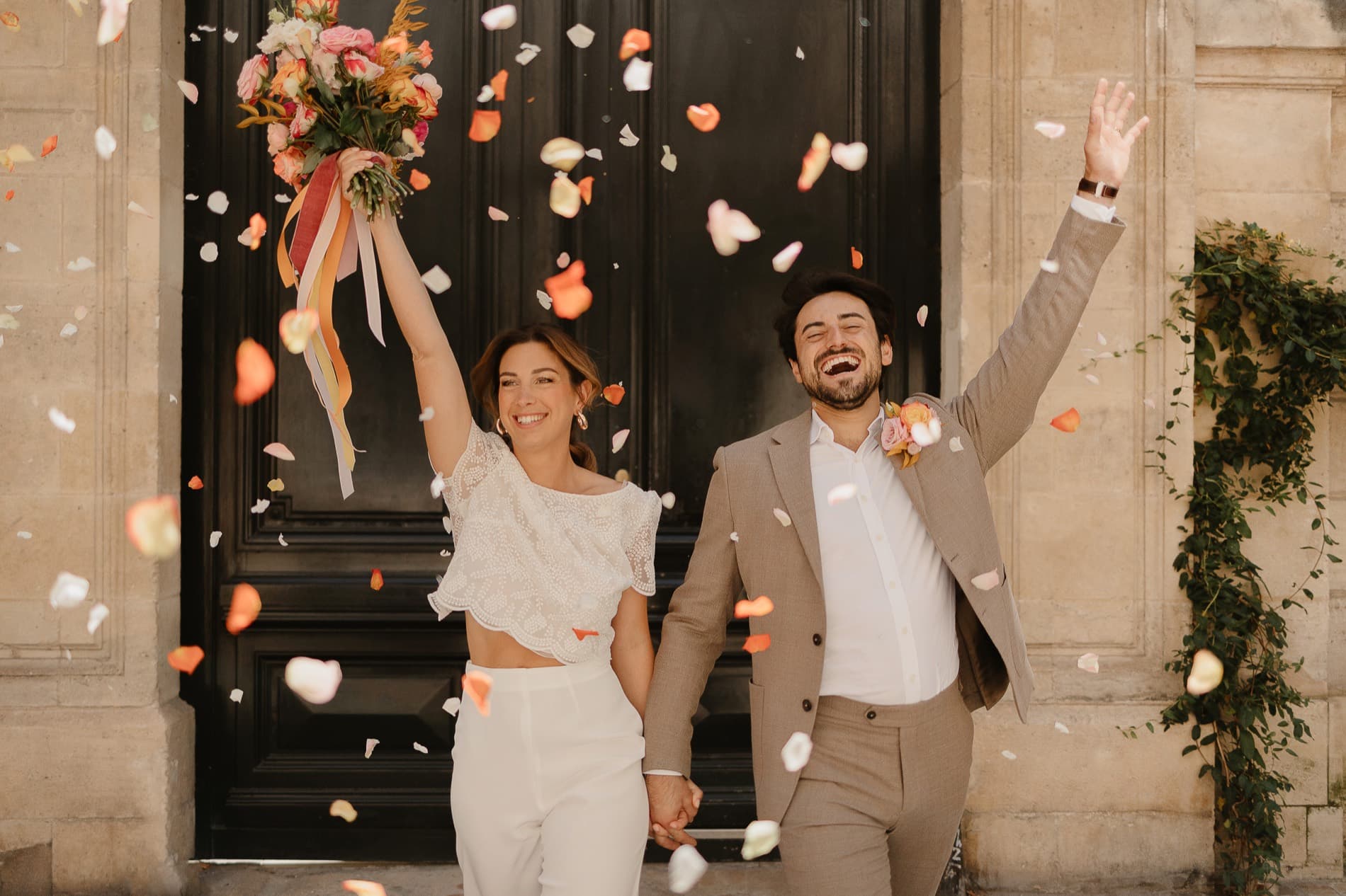 Les mariés lèvent les bras, bouquet en l’air, sous une pluie de pétales